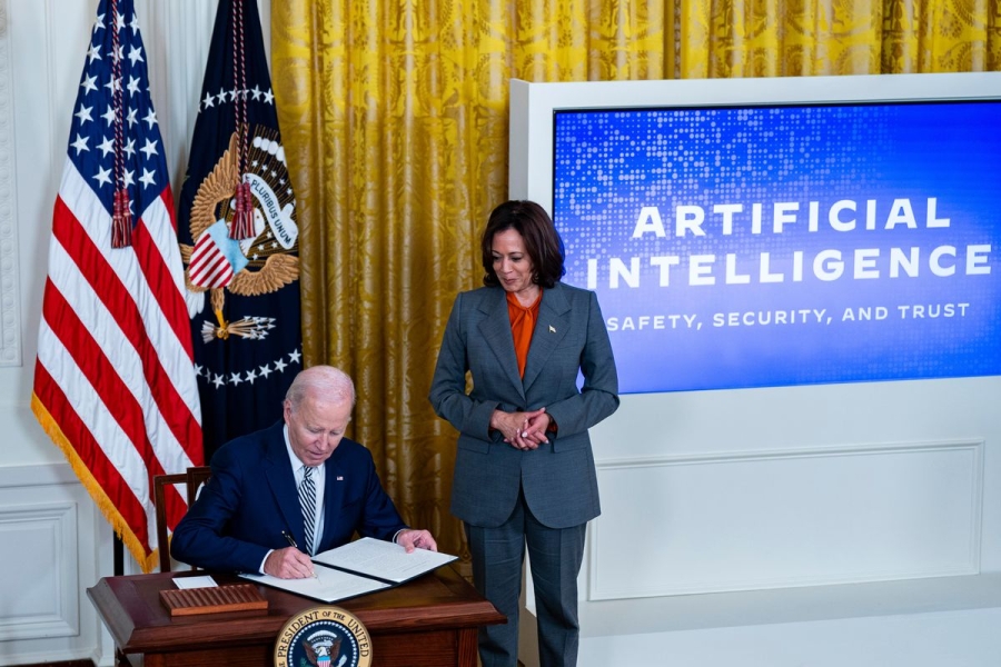 Biden sits at a desk and signs an executive order while Harris stands next to him, by a sign that says “Artificial Intelligence: Safety, Security, and Trust.”