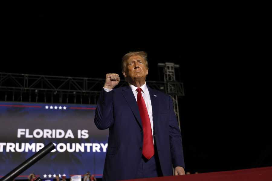 Trump wears a navy blue suit jacket and a red tie, and raises his right hand in a fist. Behind him, a screen displays the words: Florida Is Trump Country