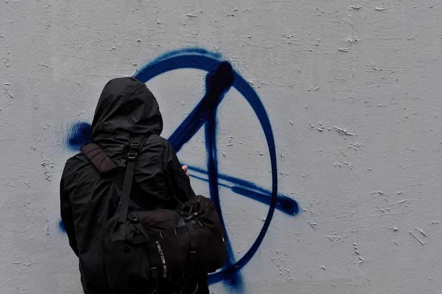 A protester, dressed in black and carrying a black bag, tags a gray wall with the anarchy sign — a capital A overlapping a circle — in blue paint.
