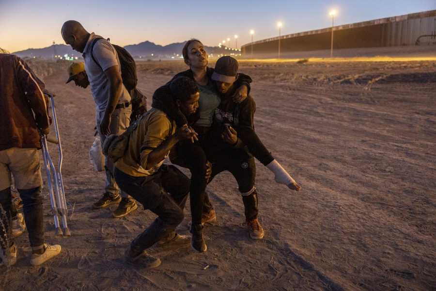 Two men carry an injured woman with a bandaged foot. Another man holds a pair of crutches. In the distance, a very tall wall-like fence, brightly lit with lamp posts, can be seen.