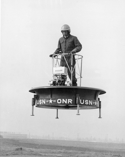 A black-and-white photo of a man in a jumpsuit and helmet standing on a circular platform that hovers in midair. 
