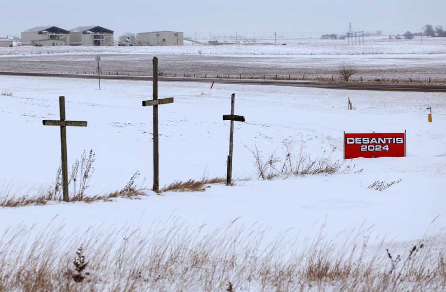 A sign reading “DeSantis 2024” is seen in a snowy field beside a road and three makeshift wooden crosses.