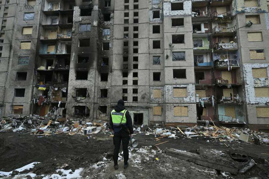 A person in a hoodie and high-visibility vest faces away from the camera and toward a destroyed building.