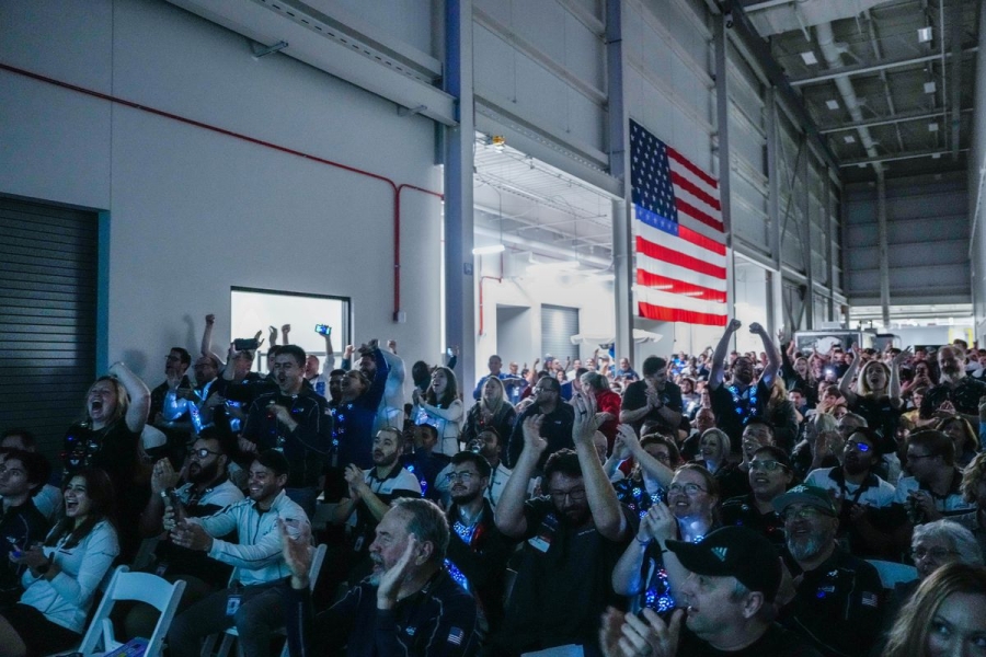 A hangar-sized room with an American flag on one wall and a crowd of cheering people.