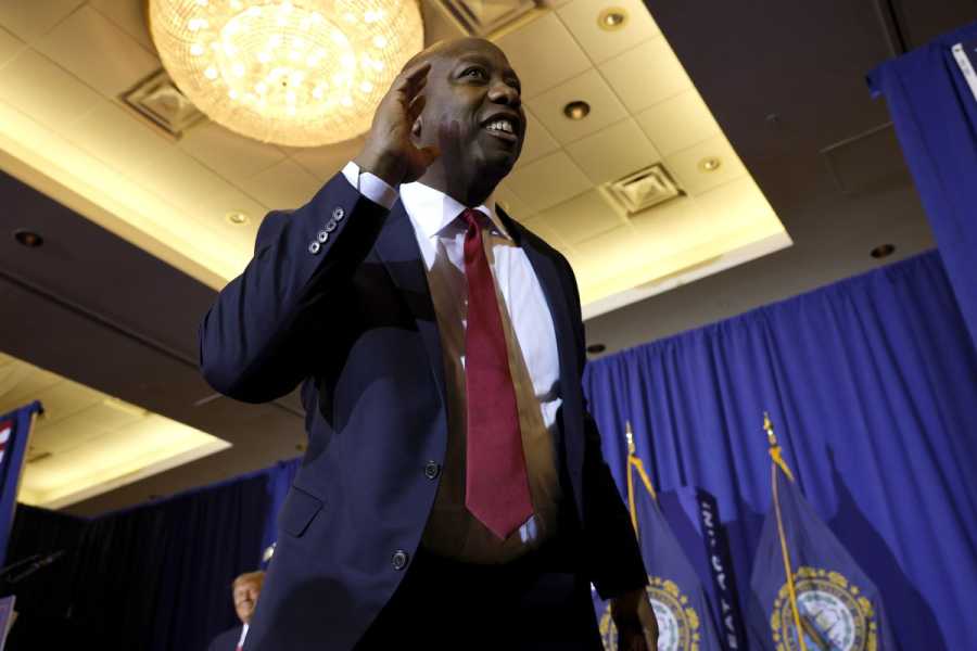 Tim Scott smiles and waves, wearing a navy suit and red tie. Blue curtains hang from the walls behind him. 