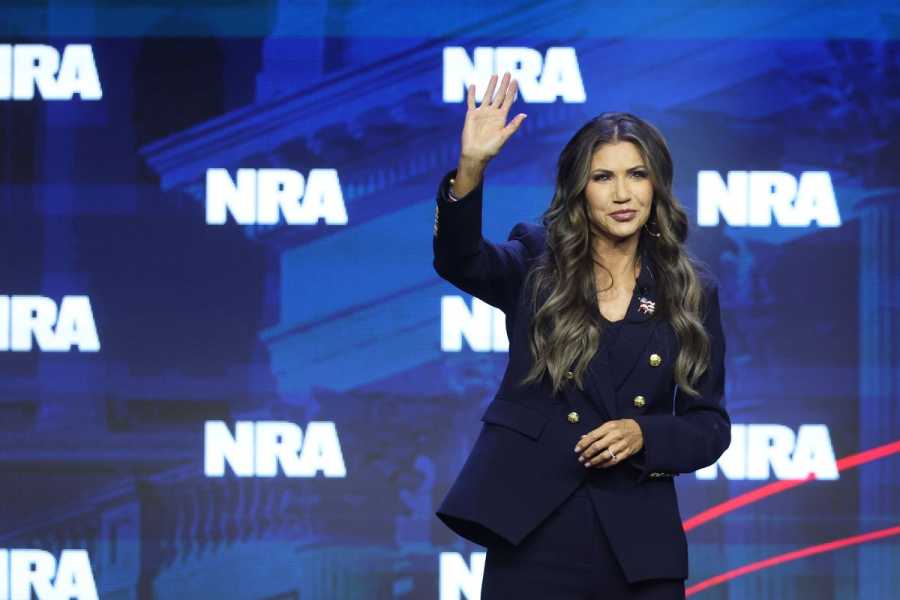 Kristi Norm waves to the audience from a stage dressed with an “NRA” backdrop.