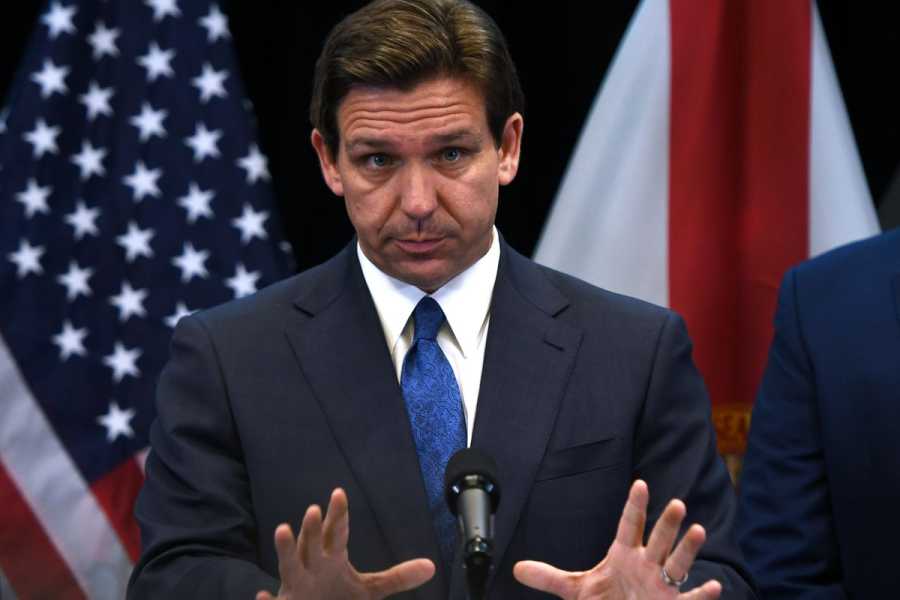 DeSantis gestures with both hands while holding a press conference, standing in front of a US flag.