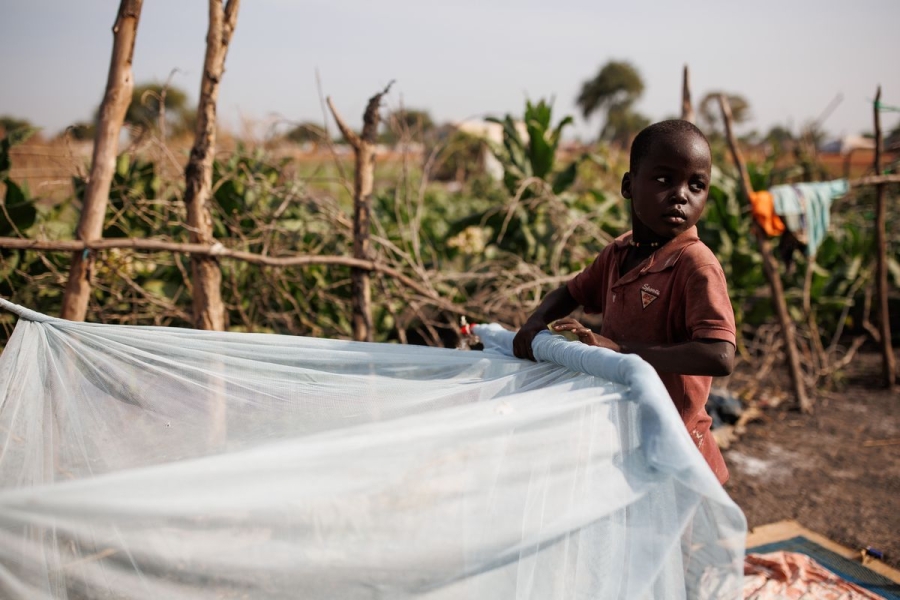 A young boy wearing a red shirt rolls up a white mosquito net covering bedding at a South Sudan refugee camp. Behind him is a makeshift fence of sticks separating the sleeping area from nearby bushes.
