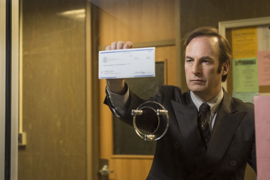 A still from the TV show Better Call Saul shows actor Bob Odenkirk in character, wearing a bad suit and pressing a check against the glass of a bank teller window. 