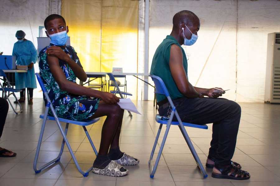 Two people wearing breathing masks sitting in folding chairs in a line in a clinic.