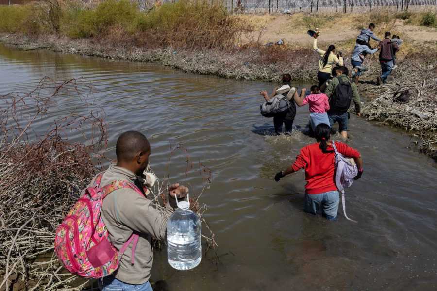 Men, women, and children carrying backpacks and bags wade through a waist-deep stream to reach the other side, forming a line of people.