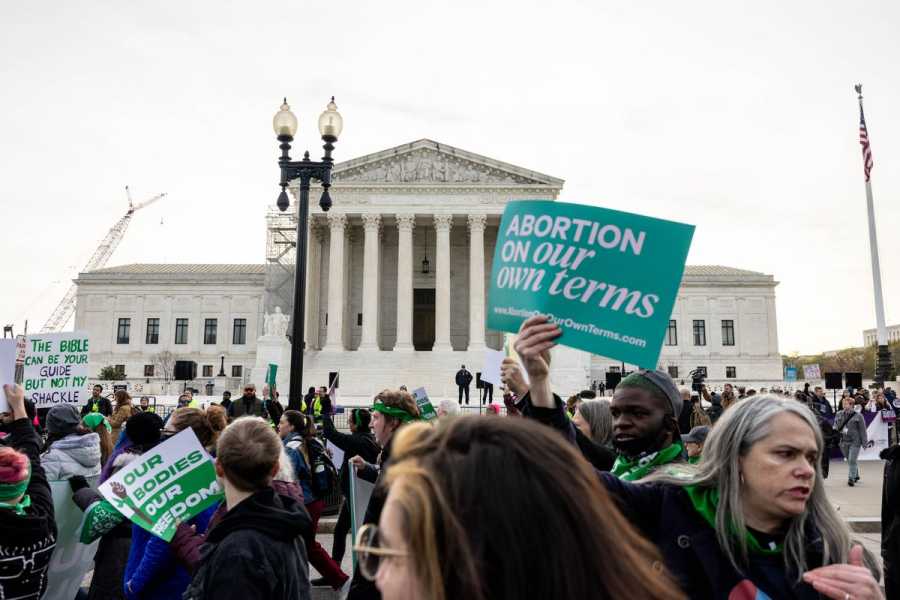 In Washington, DC, crowds gather in front of the Supreme Court building, a white marble building with columns. One demonstrator holds a blue sign that reads, “Abortion on our own terms.”