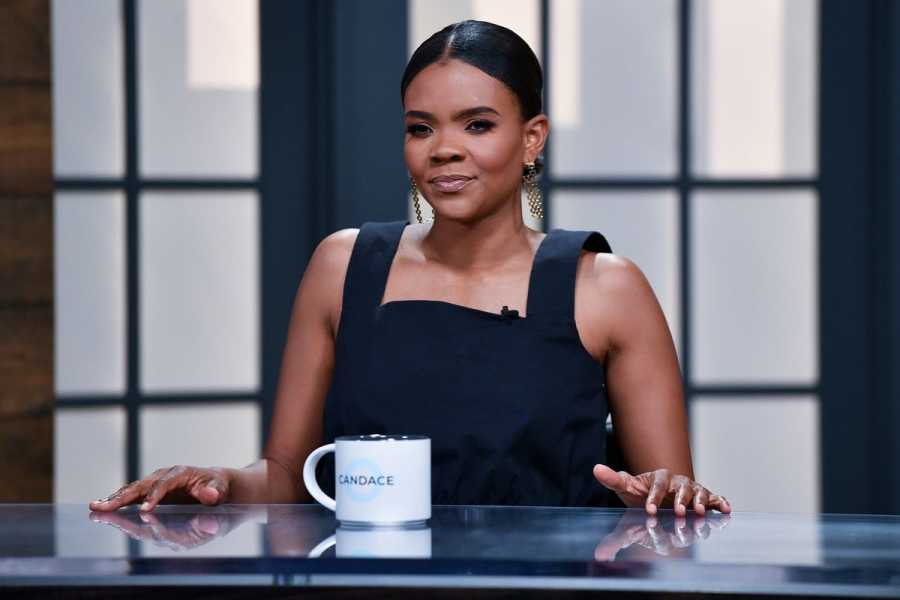 Candace Owens, a Black woman in a black square-necked sleeveless dress with her hair pulled back, sits at a glass desk with a mug reading “Candace” in front of her, and a window behind her.