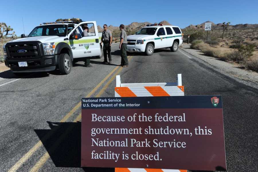 Rangers stand near two official vehicles blocking a road. A sign reads, “Because of the federal government shutdown, this National Park Service facility is closed.”