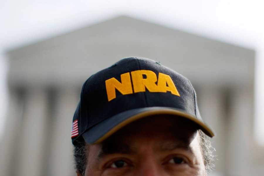 A close-up photo of a man wearing a navy baseball cap embroidered with the letters NRA in yellow. The US Supreme Court building is visible in the background.