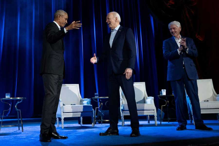 Obama, Biden, and Clinton, wearing suits, stand on a blue curtained and carpeted stage on which three white armchairs are arranged.
