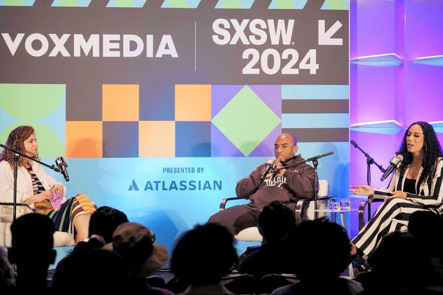 Three people sit in chairs with microphones on an event stage at SXSW. 