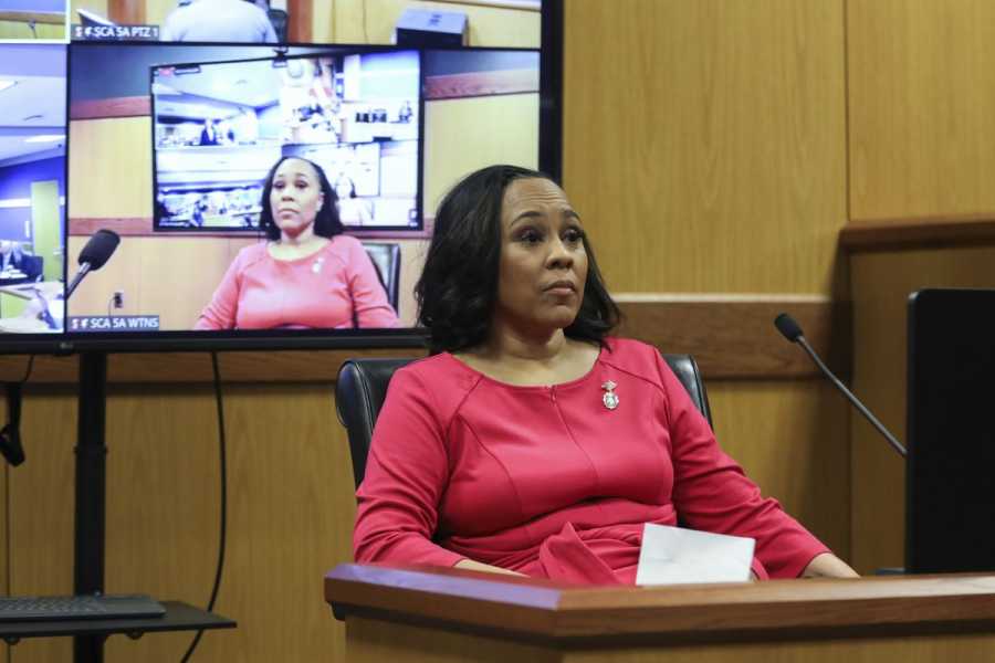 A Black woman with long straight hair wearing a red long-sleeved dress sits at a courtroom table, with a video monitor behind her showing her testimony. 
