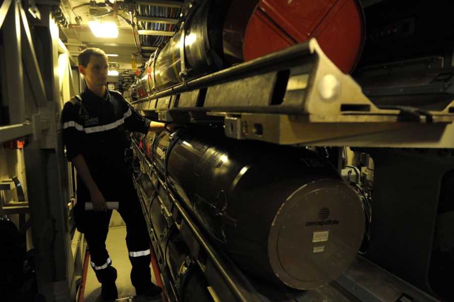 A uniformed crew member inspects torpedoes inside a dimly lit submarine.