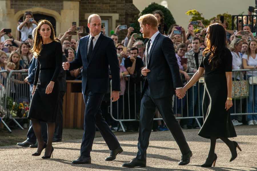 Princess Catherine, Prince William, Prince Harry, and Meghan, Duchess of Sussex, walk side by side, all dressed in black. A crowd of people watch them from behind a barricade. 