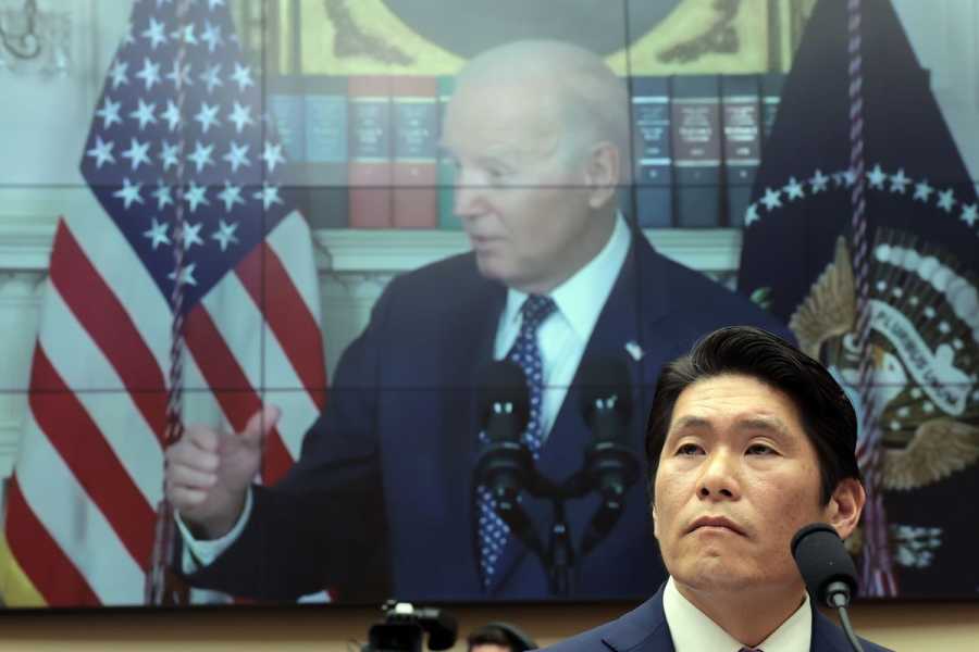 Robert Hur, an Asian man wearing a dark suit, sits at a congressional desk with a microphone in front of him and a large video screen showing Joe Biden behind him. 