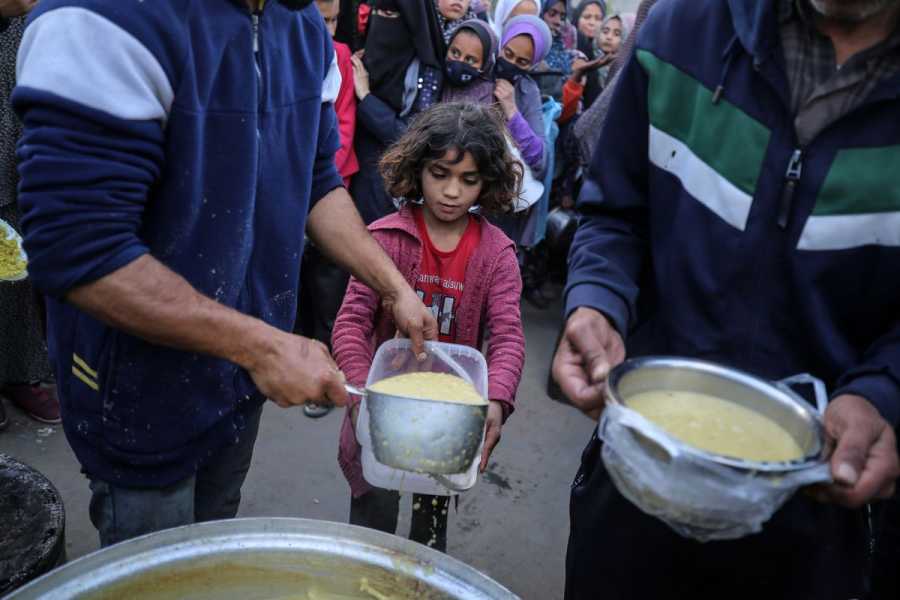 Two men, their faces not shown, ladle food from a large metal pot into a small plastic container held by a child in a pink jacket. Behind the child, many wait in line for food.