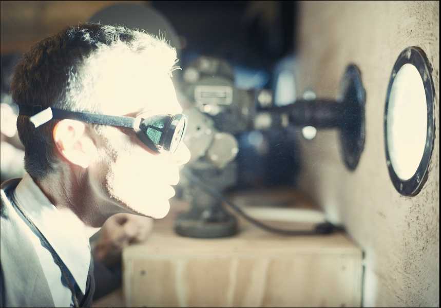 Oppenheimer wearing goggles, his face lit from the blast of the bomb as he watches through a circular window.
