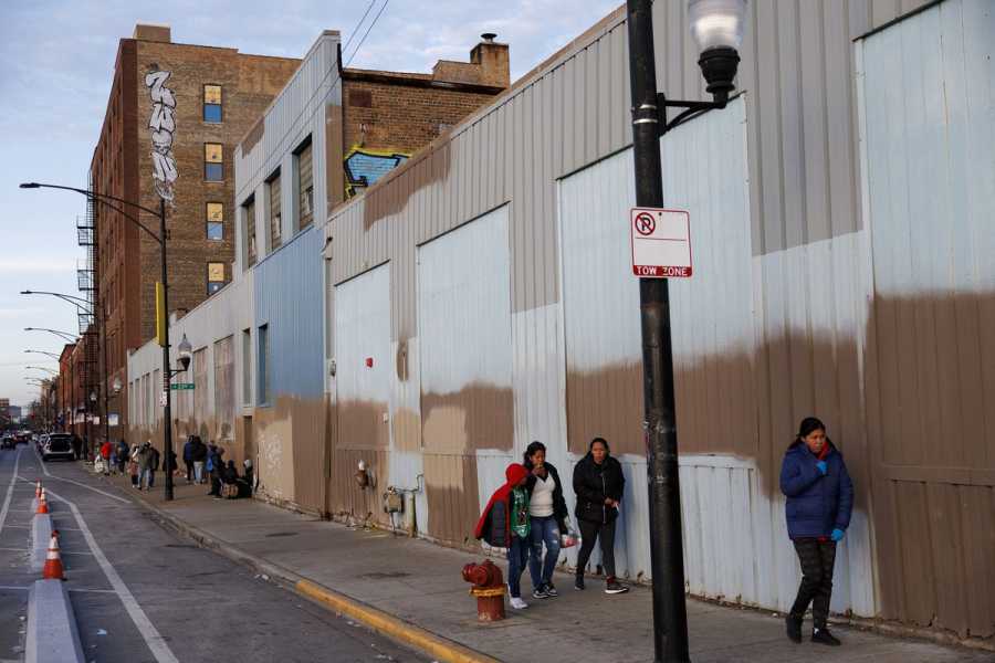Several people walk on a sidewalk flanked by a haphazardly painted warehouse. 