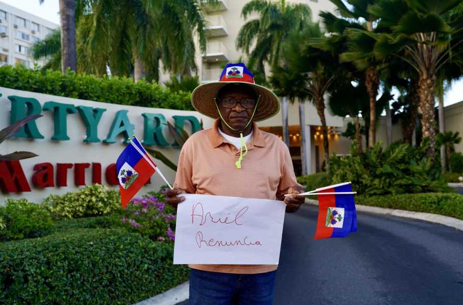 A man with a serious expression holds Haitian flags and a sign reading “Ariel renuncia.”