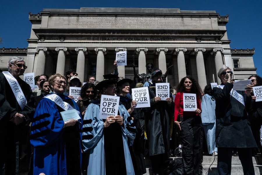 A group of faculty, many in ceremonial robes, hold signs and stand on the steps of a large building at Columbia University.
