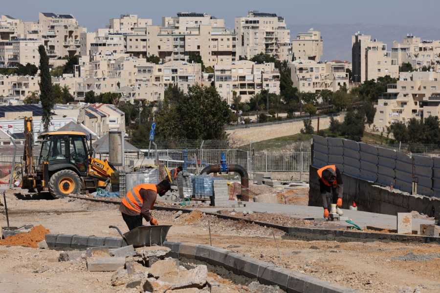 Men in orange vests at work in a dusty construction site. Multistory urban buildings made of tan concrete stand in the background.