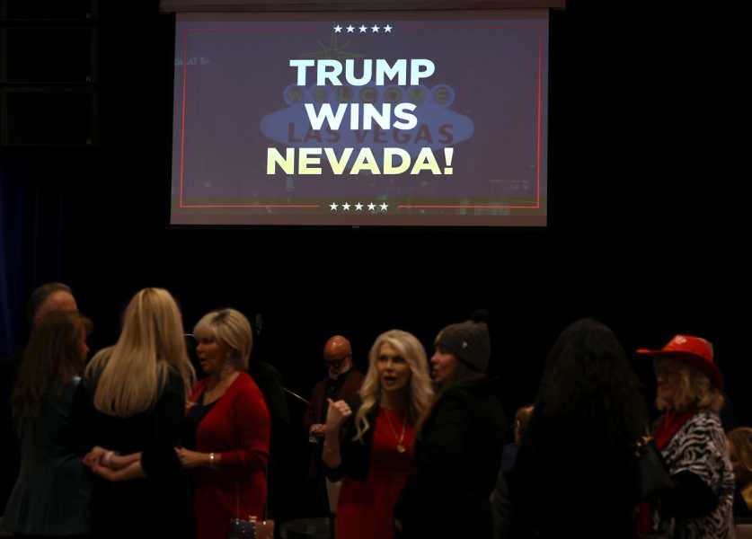 A crowd of people stand in a dimly lit room. Above them hangs a screen displaying the words: TRUMP WINS NEVADA! 