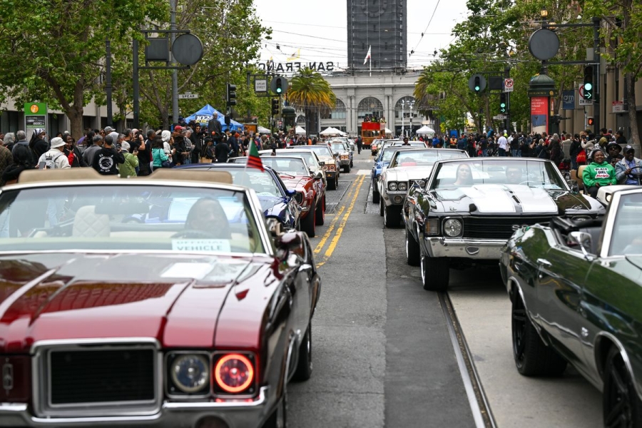 Older cars drive along a street lined with people.