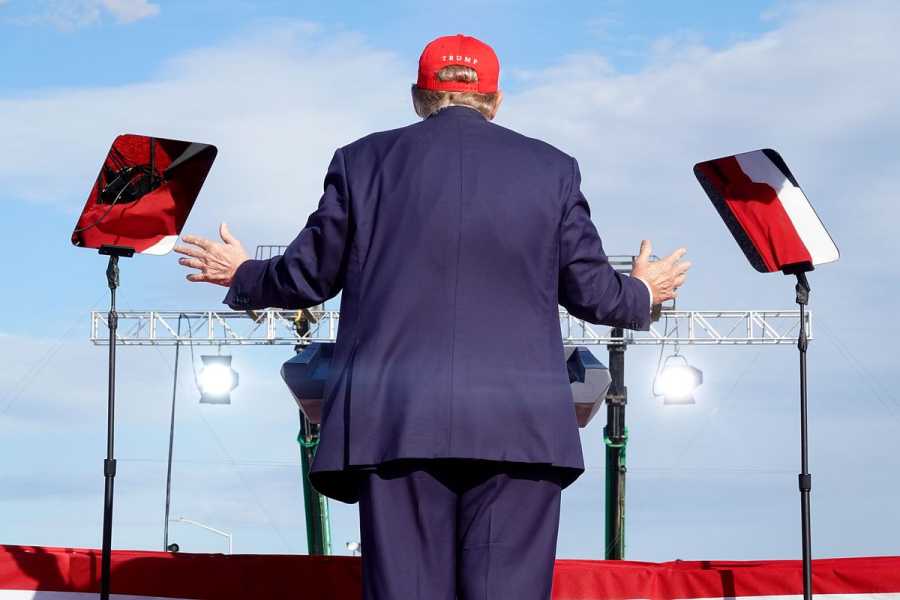 Trump, wearing a navy suit and a red baseball cap, faces away from the camera while delivering a speech.