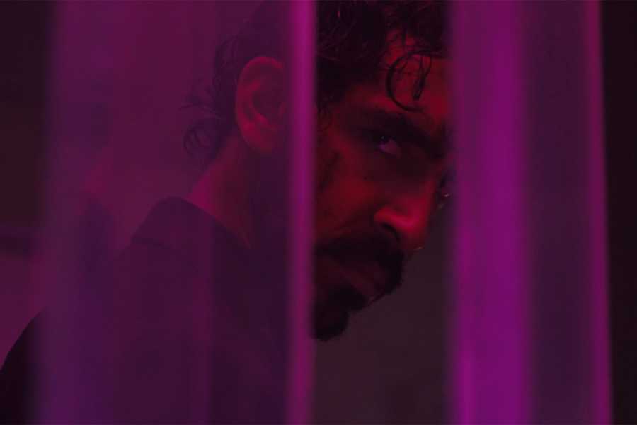 Patel, with a mustache and beard, peers between thin columns of purple glass, his face illuminated by red light.