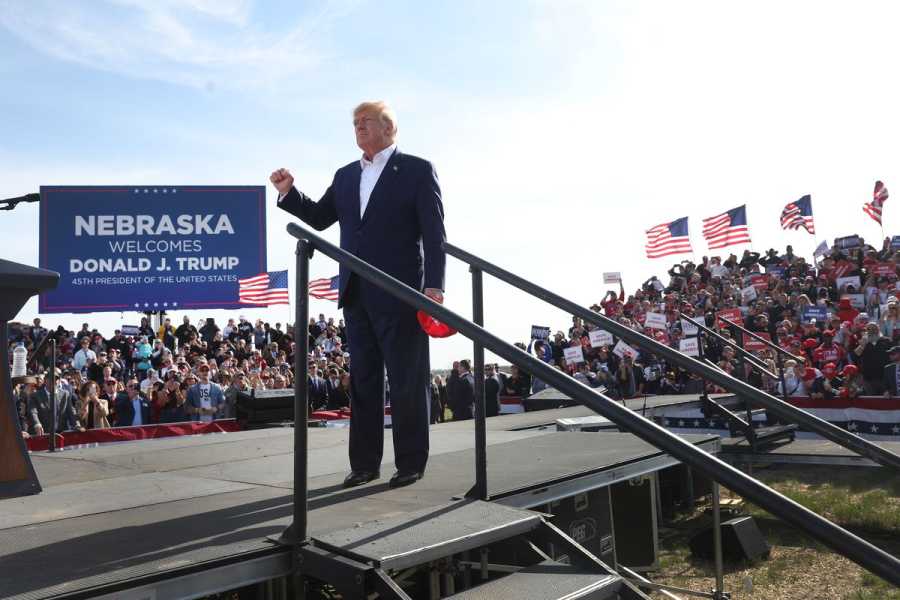 Donald Trump, in a dark suit and red tie, stands on a temporary platform surrounded by supporters with American flags and a large blue sign saying “Nebraska welcomes Donald Trump.”