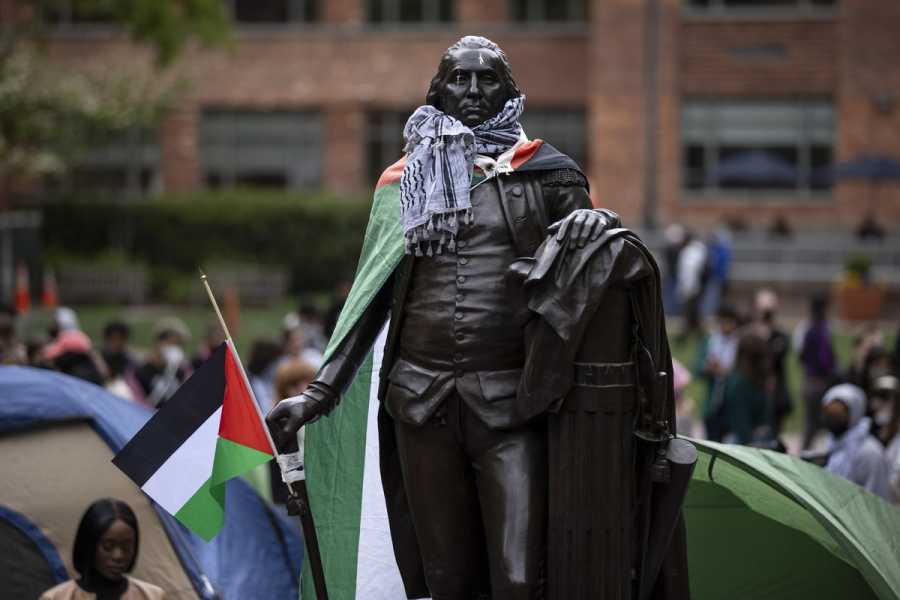 A statue of George Washington has a keffiyeh around its neck and a Palestinian flag as a cape. Behind it, students camp in tents and sit on the grass.