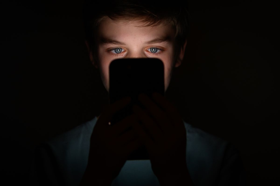 A teen in a dark room holds a phone in front of their fact. The light from the screen illuminates their eyes.