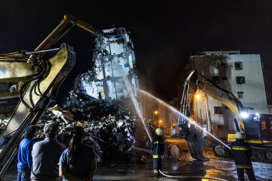 Fire and rescue crews spray water on a pile of rubble as construction equipment demolishes a multi-story building, with crowds watching from the street.