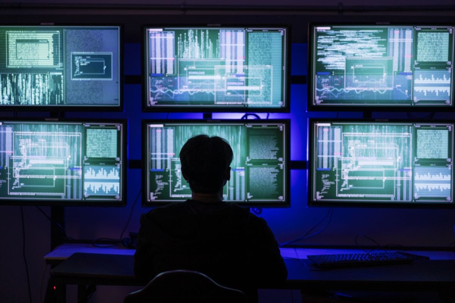 A silhouetted person sitting in front of six computer screens full of data.