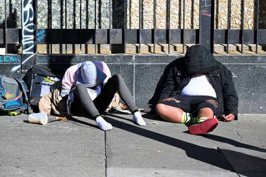 Two people in hoodies sit on a sidewalk with their backs against a low wall and their heads down.