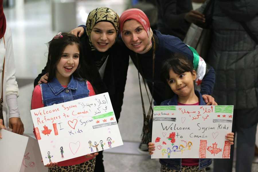 Two children and two women hold child-made illustrated signs welcoming Syrian refuges. One sign reads “Welcome Syrian refugees to Canada, we (heart) Syrian kids,” the other reads “Welcome Syrian kids to Canada.” Both signs are also written in Arabic.