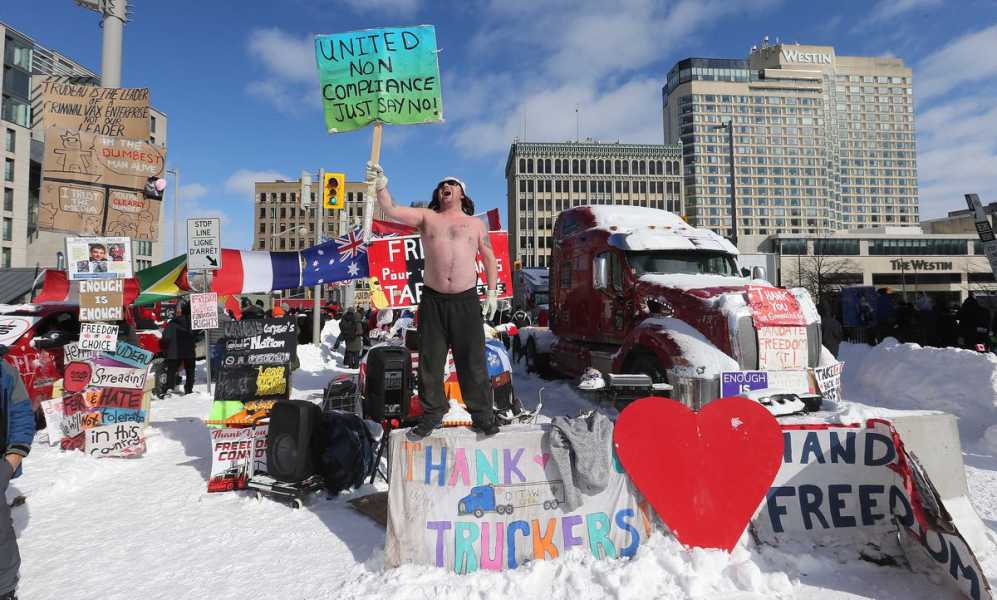 A shirtless man carrying a sign that reads “United non compliance: just say no!” stands atop a pile of gear that sits beside a semi truck blocking a road in downtown Ottawa, Canada.