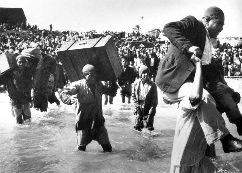 A black-and-white photo of large crowds wading into water carrying large suitcases on their heads and shoulders. One man carries another man on his shoulders. 