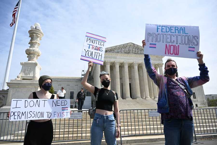 People stand holding signs in front of the Supreme Court building, including “Protect Trans Youth.”