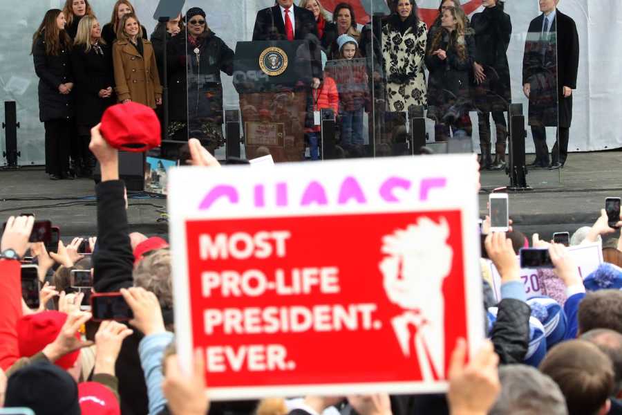 A view of Donald Trump speaking on a temporary platform in front of a large crowd holding signs including “Most Pro-Life President Ever.”