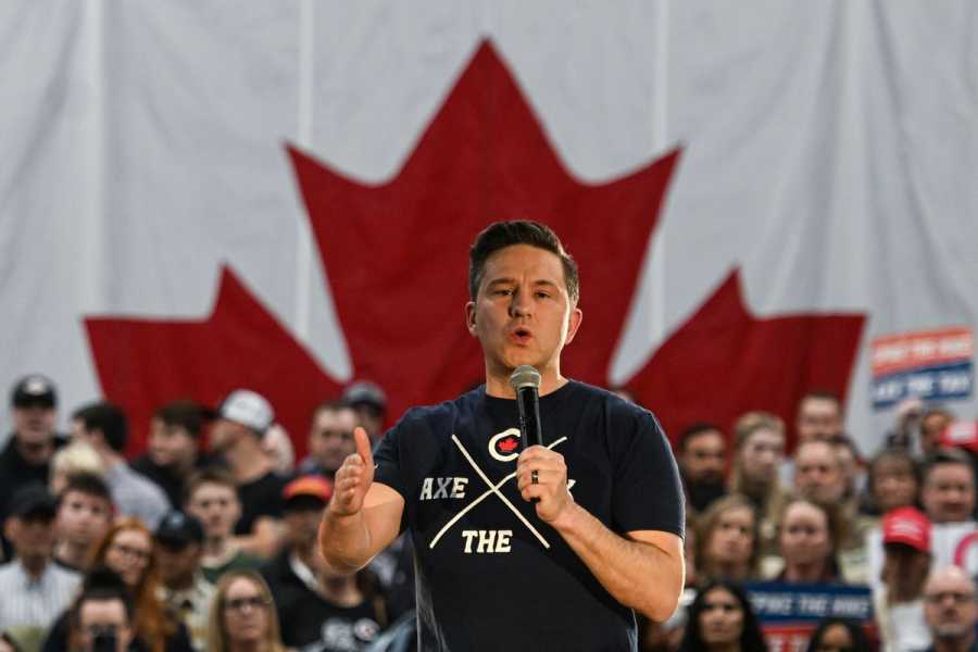 A man in a black T-shirt speaking into a handheld microphone in front of a crowd and a Canadian flag backdrop.