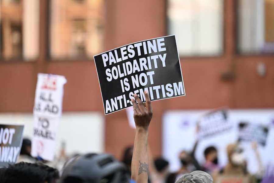 One person within a crowd of people in front of a school building holds a sign that reads “Palestine solidarity is not anti-semitism.”