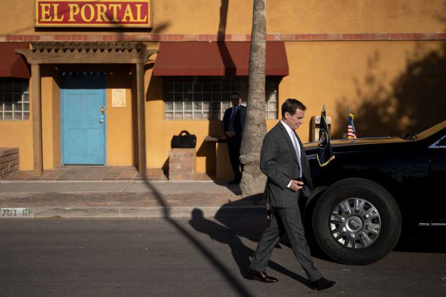 John Kirby, wearing a suit, walks past a black SUV flying a small American flag and official government flag. Behind him is a yellow building with rust-colored awnings and a blue door. 