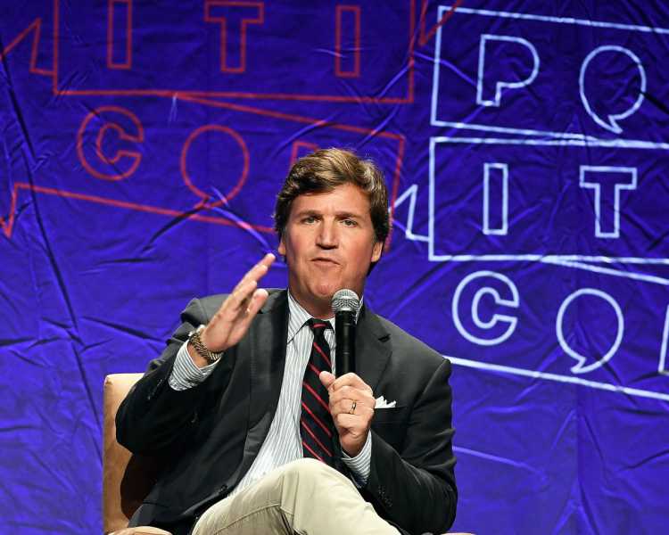 Carlson, sitting cross-legged onstage at Politicon 2018, gestures while speaking into a microphone.